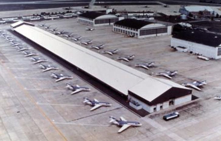 Aerial view of a long hangar and older hangars with small jets parked on the apron