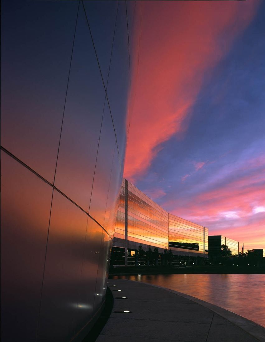 FedEx: Curved metal wall beside a waterside path, with a long glass building reflecting a pink sunset