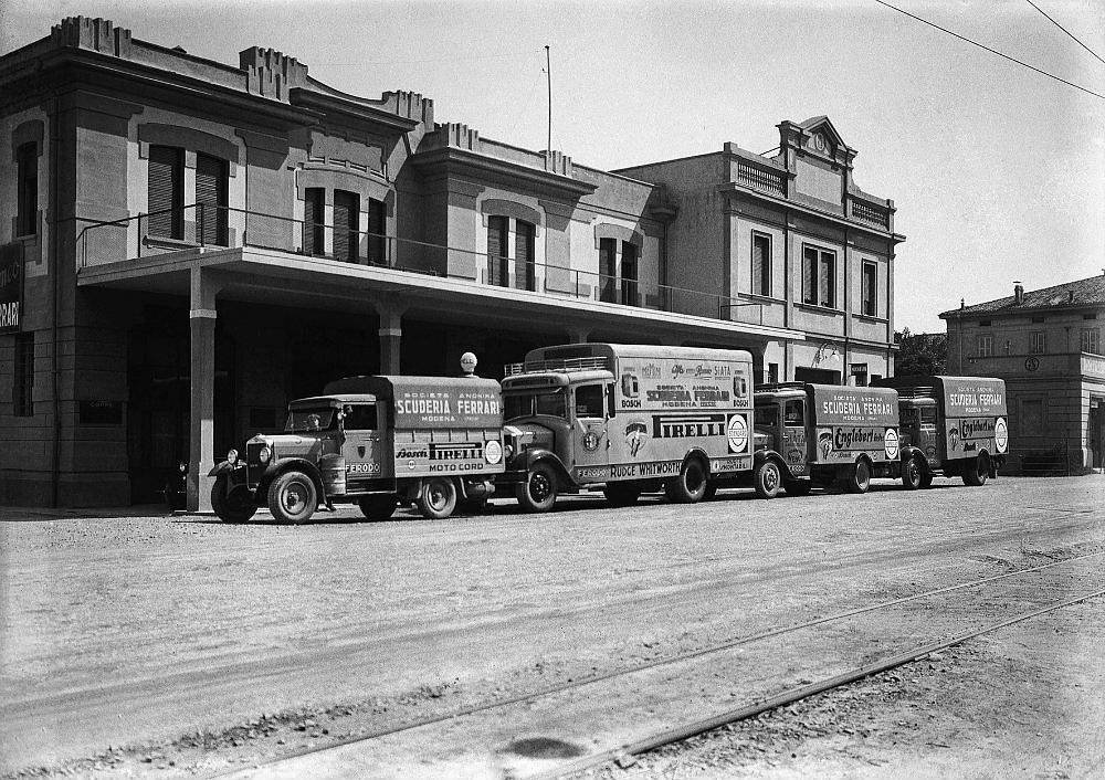 Black-and-white photo of Scuderia Ferrari trucks outside a building