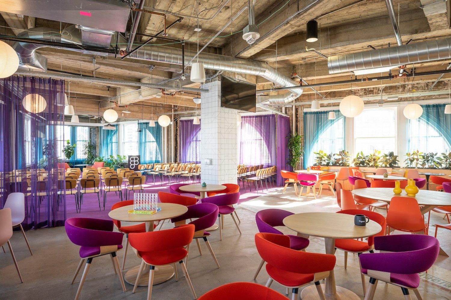 Open lounge with round tables, red and purple chairs, exposed ceiling ducts and tall windows