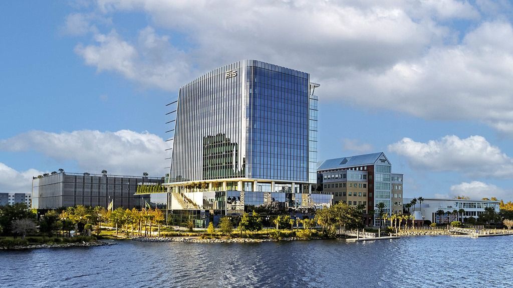 A glass office tower on a river bank under a blue sky with a riverside park in front