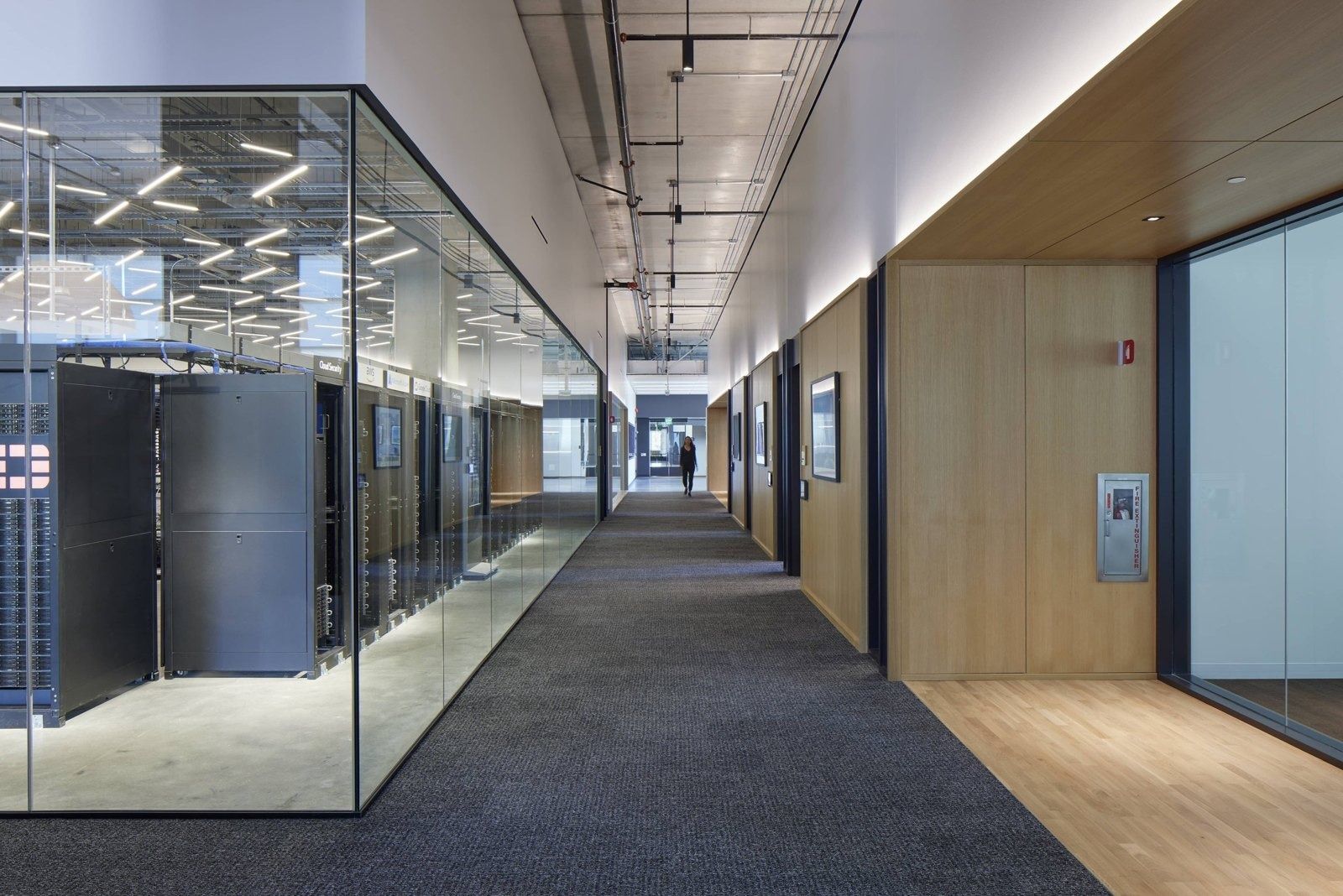 Fortinet: Corridor with dark carpet beside a glass-walled room holding lit equipment racks, with wood-paneled walls opposite