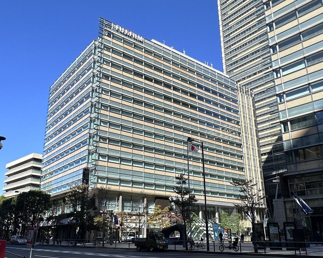 Glass-clad mid-rise office building with horizontal louvres under a blue sky, a taller tower beside it