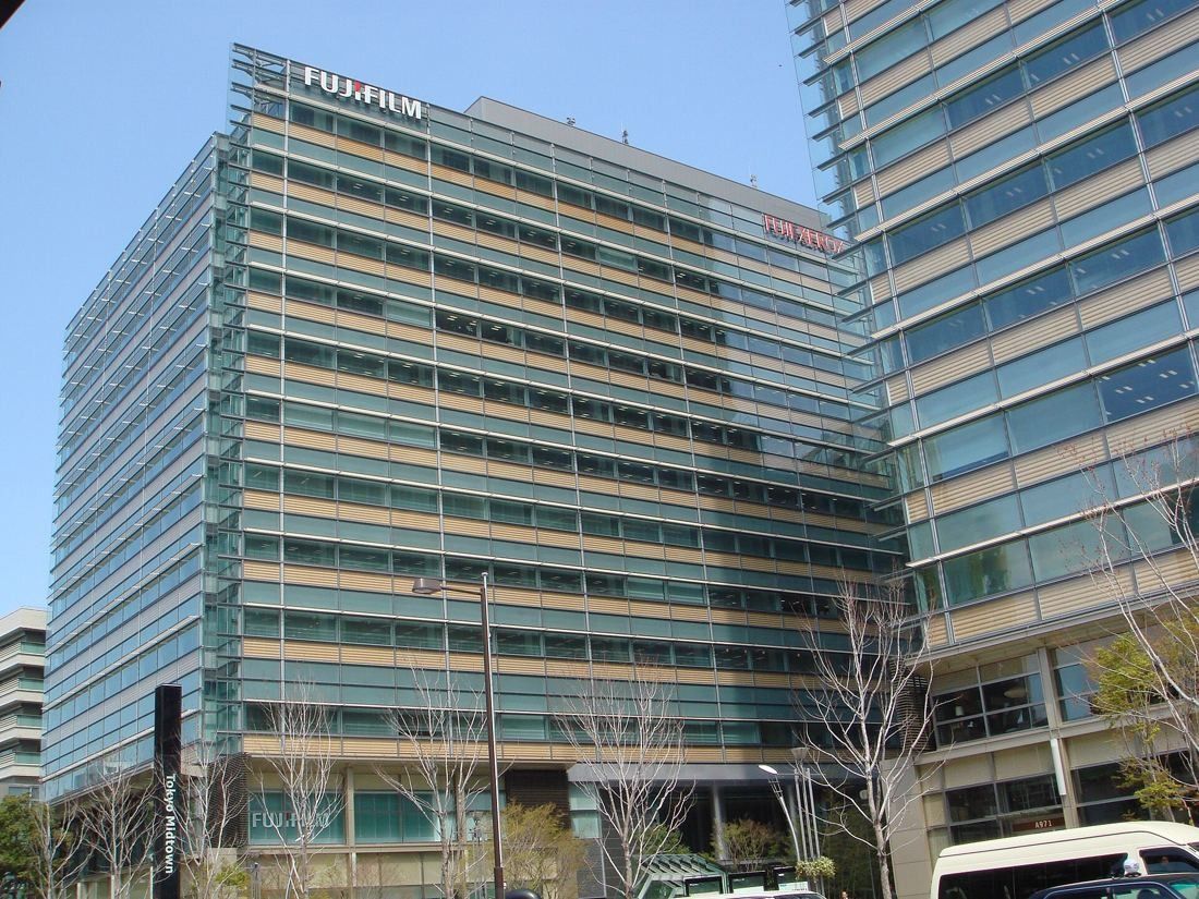Fujifilm: Wide view of a mid-rise office building with a tower beside it and bare trees along the street