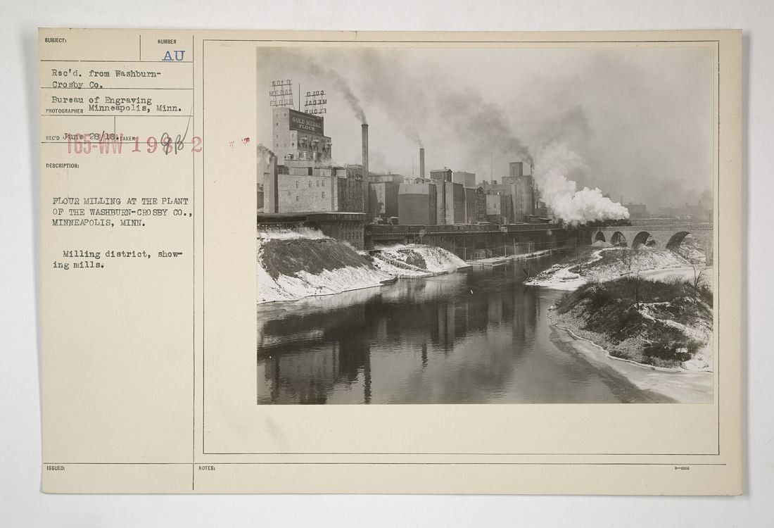 Black-and-white view along the river of a row of large mill buildings with smoking chimneys