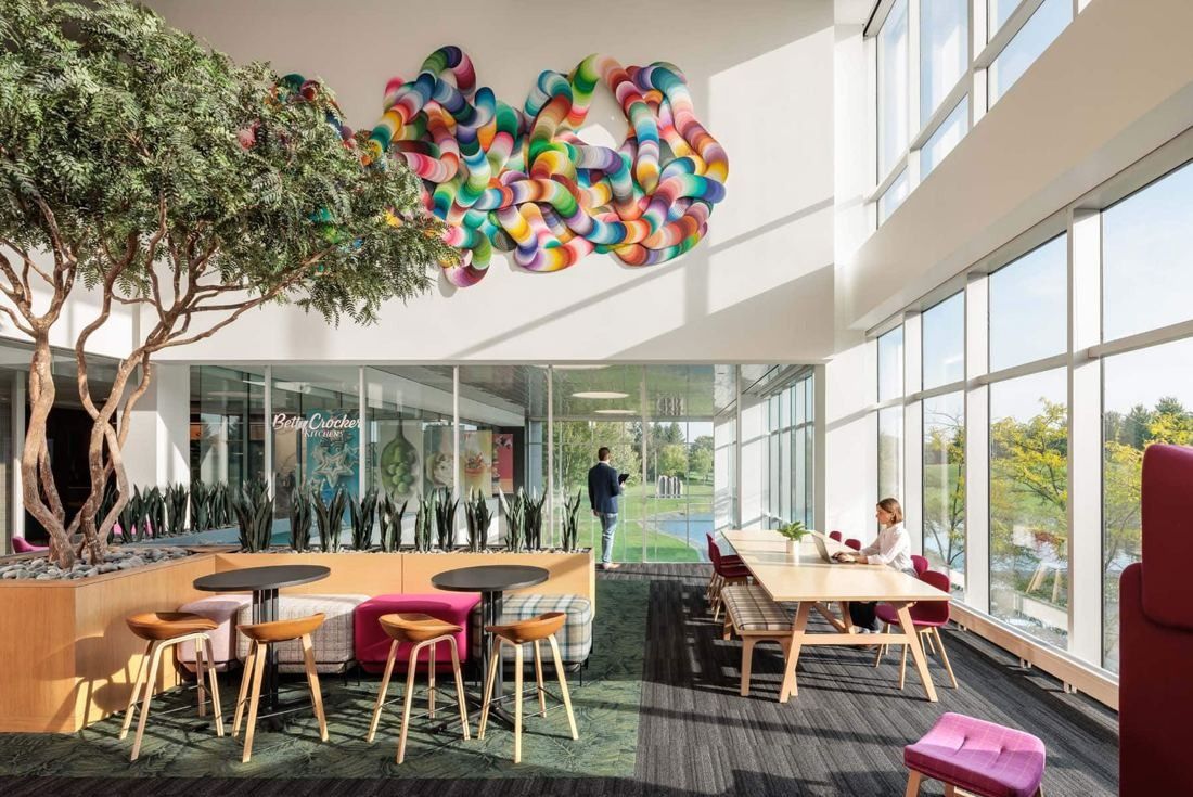 Double-height lounge with a planted tree, a colorful looped wall sculpture and tall windows onto the grounds