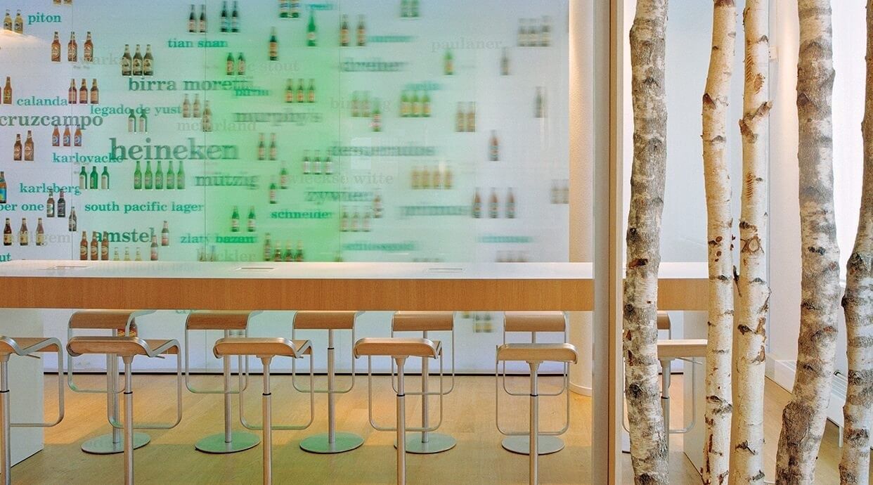 Long bar counter with stools in front of a wall printed with beer bottles and brand names, beside a screen of birch trunks