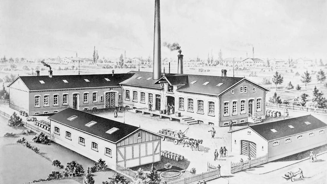 Engraving of a factory yard with low buildings and a tall chimney
