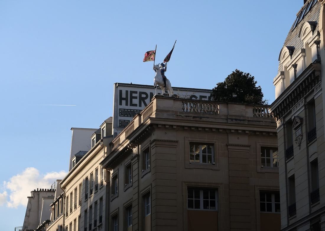 Hermès: Top of a stone corner building with a rider statue holding flags and a large sign