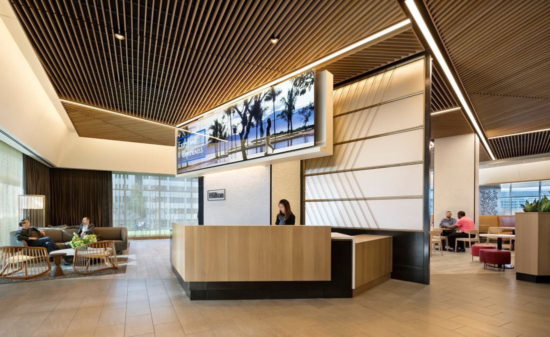 Hilton: Lounge with a wood desk, a wide screen above and armchairs by tall windows
