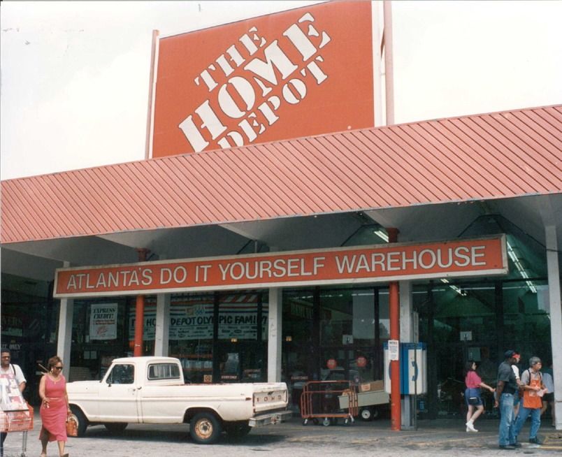 Store entrance with a large orange 'The Home Depot' sign and an 'Atlanta's Do It Yourself Warehouse' banner, shoppers and a white pickup truck in front