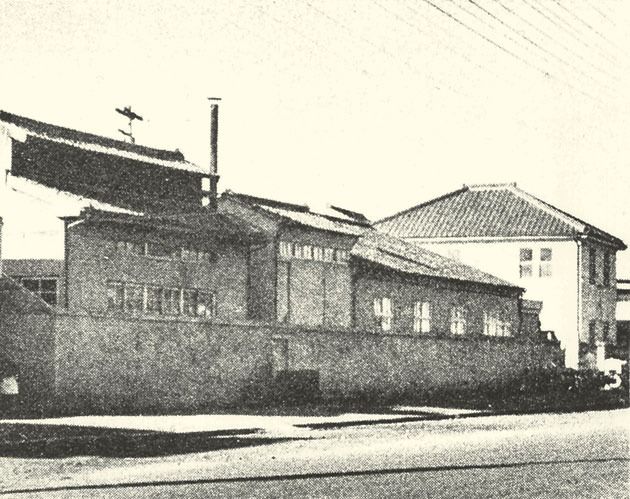Black-and-white photo of low tiled-roof factory buildings behind a wall on a street corner
