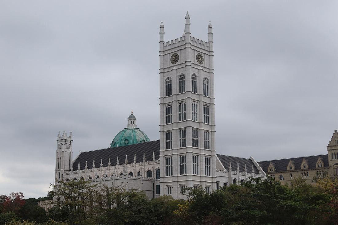 Huawei: Pale stone Gothic-style tower beside a domed hall, seen across greenery