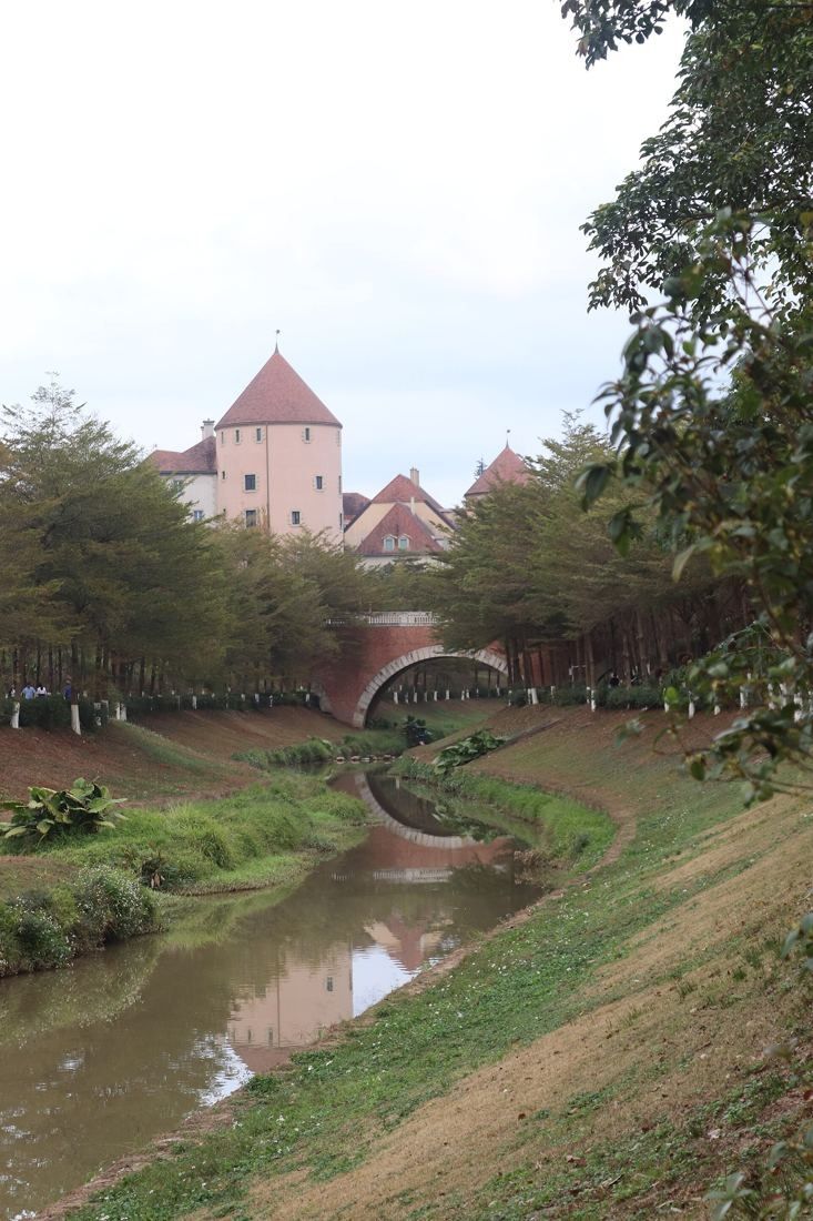 Huawei: Small stream with a stone bridge and a pink turreted building behind trees
