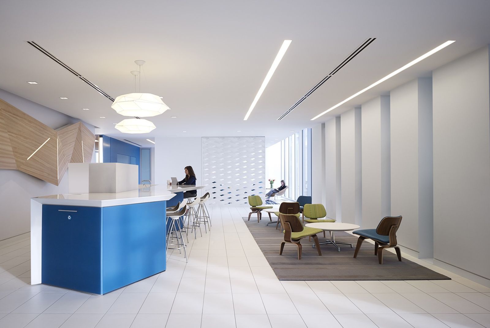A bright open office with a blue reception counter, pendant lamps and a lounge by tall windows