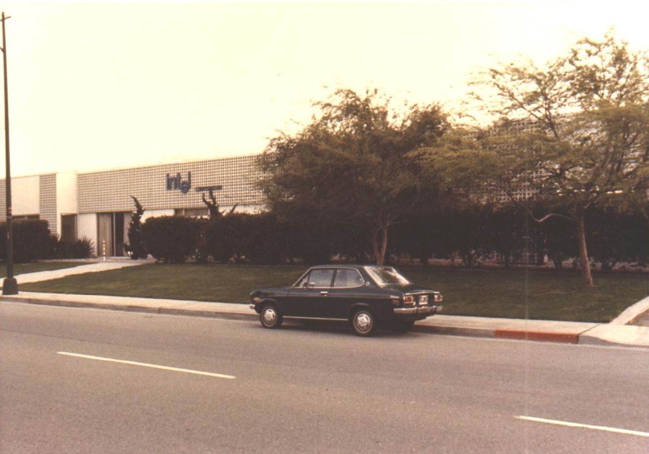 Low pale building with an Intel sign behind lawn and trees, a dark sedan parked on the street in front
