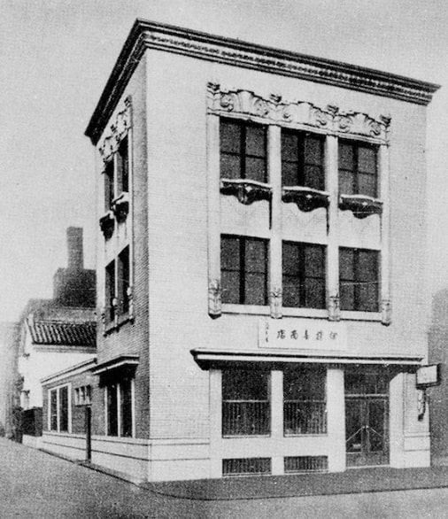 Black-and-white photo of a three-storey corner building with ornamented upper windows