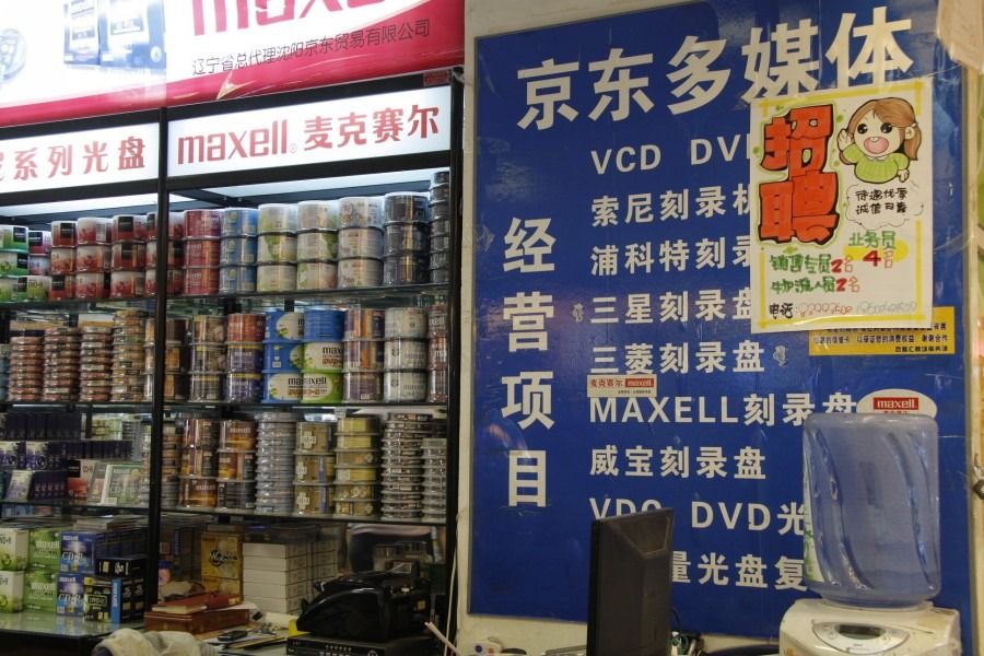A small shop counter with shelves of blank CDs and a blue sign reading “Jingdong Multimedia” in Chinese
