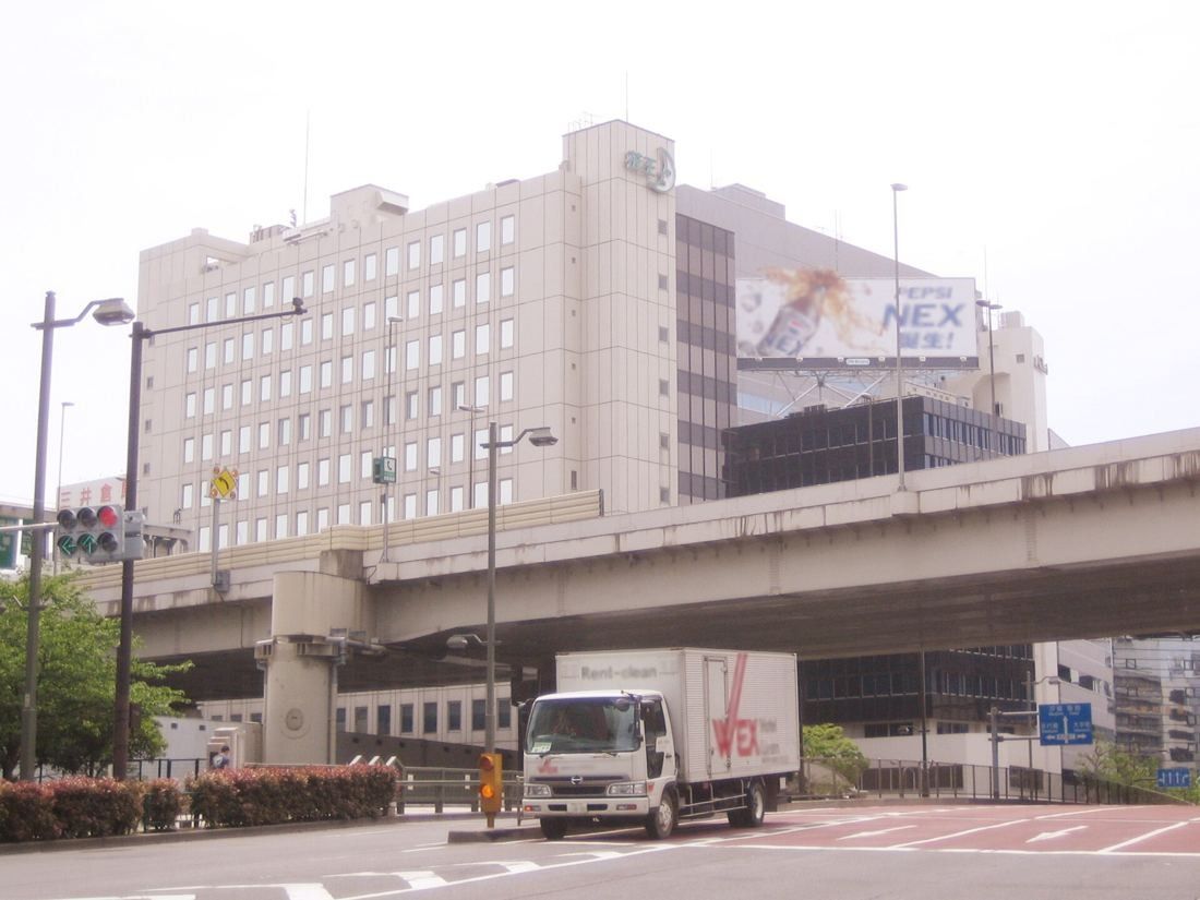 Kao: A pale office building behind an elevated expressway, with a truck on the road in front
