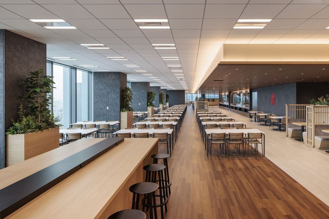 KDDI: A long dining hall with a wooden counter and rows of tables stretching toward the windows