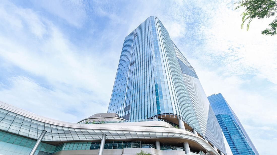 A tall glass tower rising above a curved, multi-level podium under a blue sky with clouds