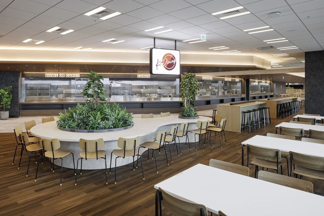 KDDI: A wide dining hall with wooden floors, round tables around a planted island and a buffet counter at the back