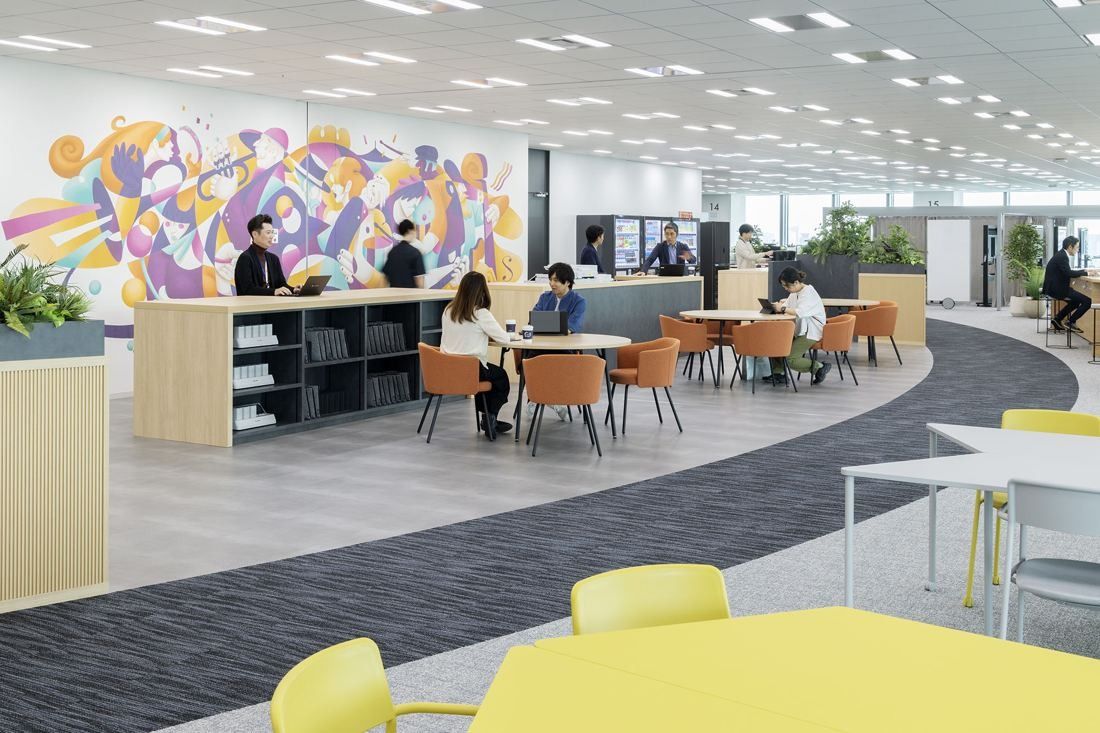 KDDI: An open work area with a colourful wall mural, orange chairs around round tables and a curving carpet path