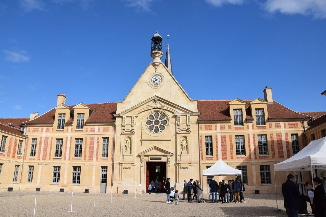 Kering: Chapel facade with clock and bell tower facing a sunlit courtyard, a few distant visitors