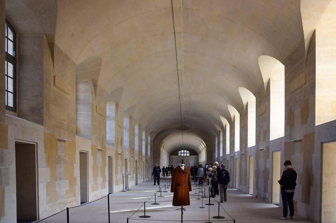 Kering: Long vaulted stone gallery lit by tall side windows, with a coat displayed on a mannequin