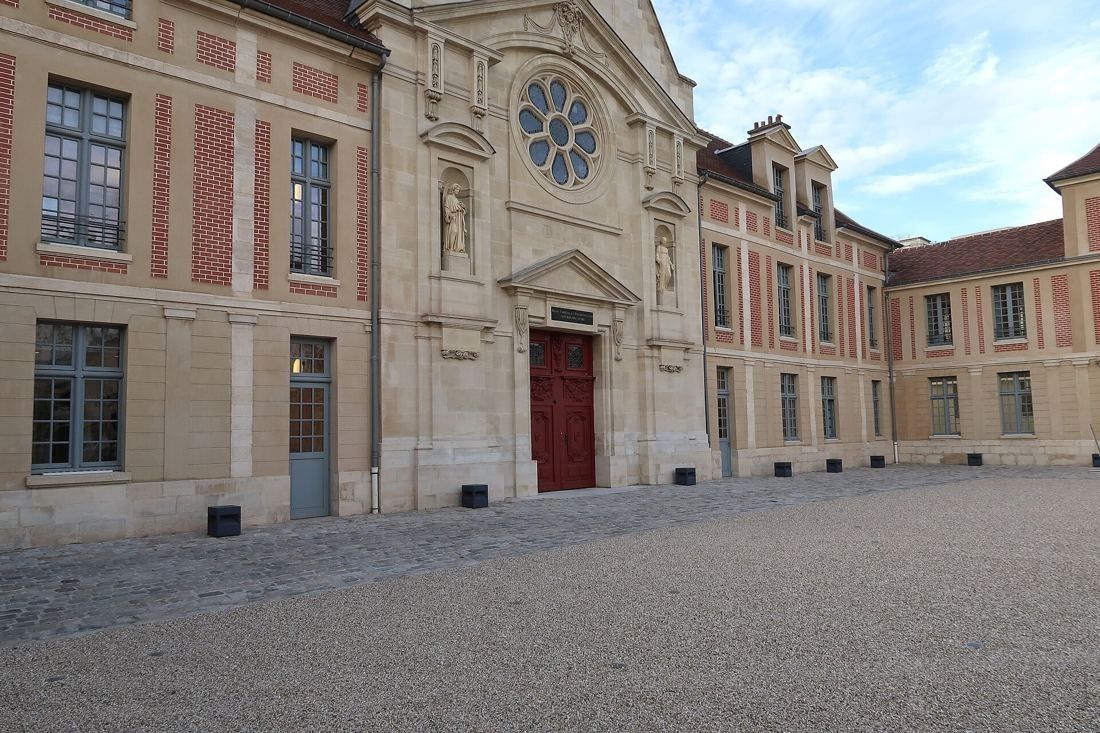 Kering: Paved courtyard in front of a chapel facade with a rose window and red door