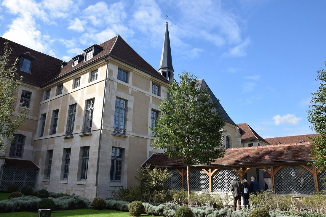 Kering: Stone wing with a slate spire behind and a timber-framed lattice gallery in a planted courtyard