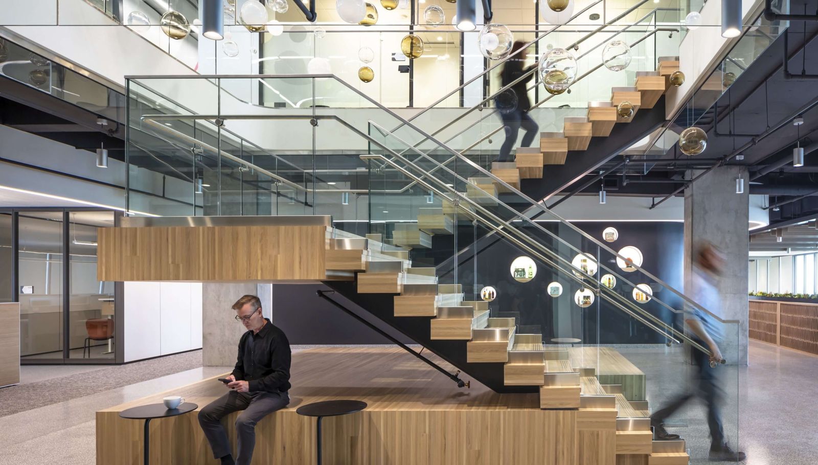 A wide timber-and-glass stair with people sitting on the steps under hanging glass globes