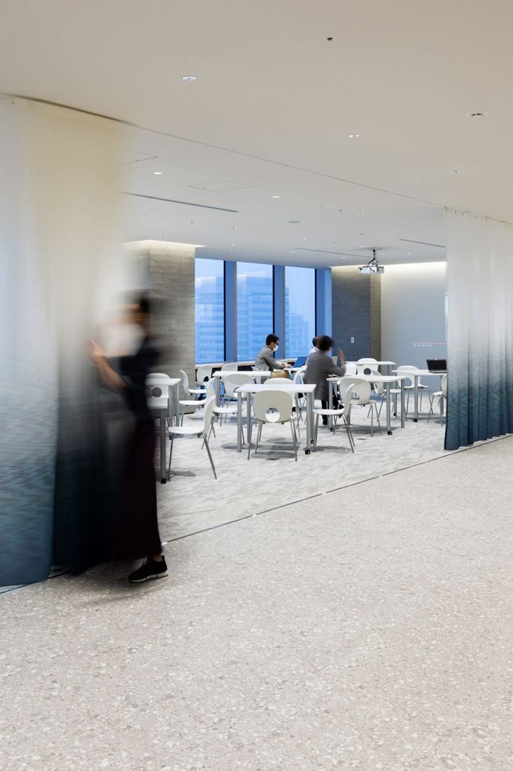 Kirin Holdings: Corridor opening onto a bright meeting area with white tables, a blurred figure walking past