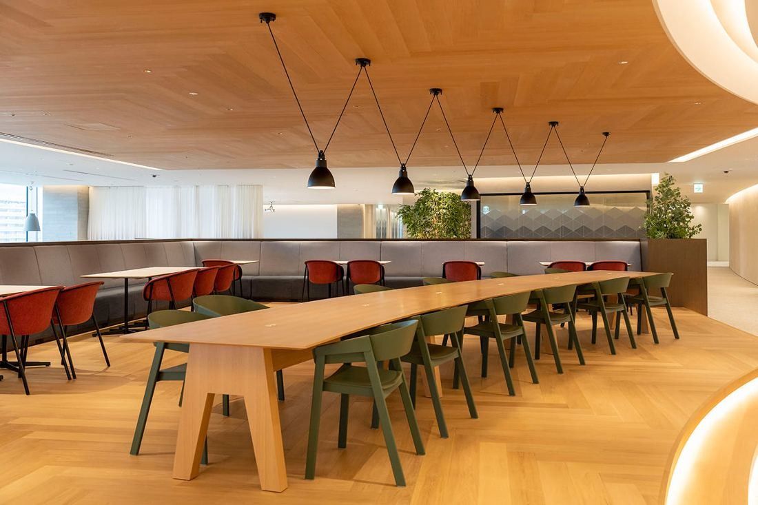 Kirin Holdings: Long wooden communal table with green chairs under black pendant lamps