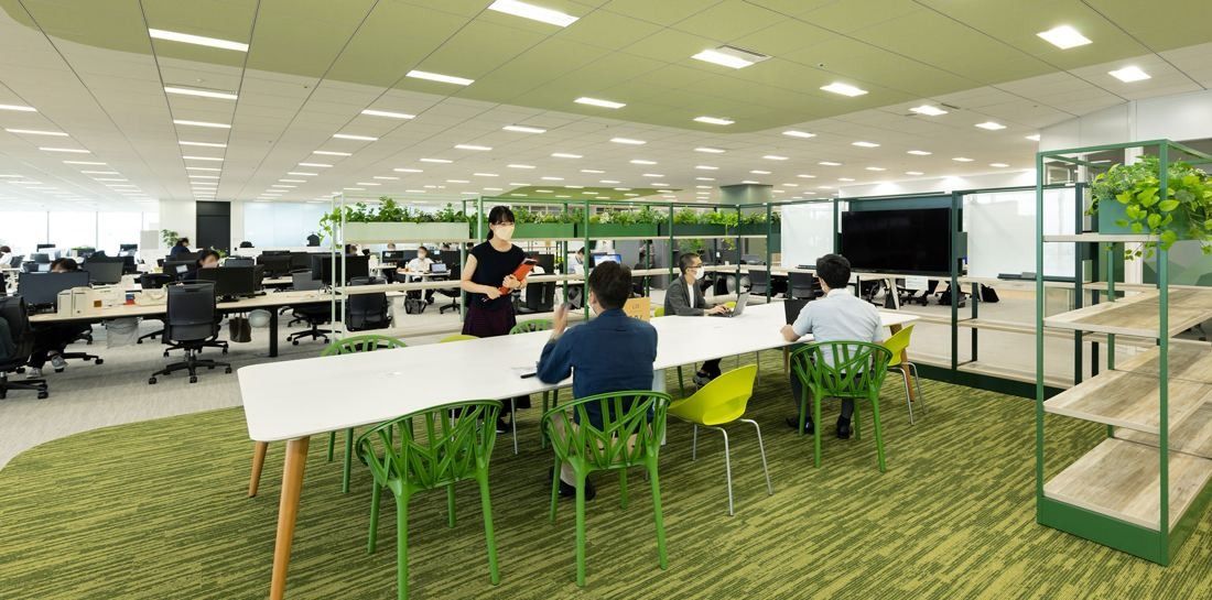 Kirin Holdings: Office floor with a green carpet zone, long white tables with green chairs and plant-topped shelving