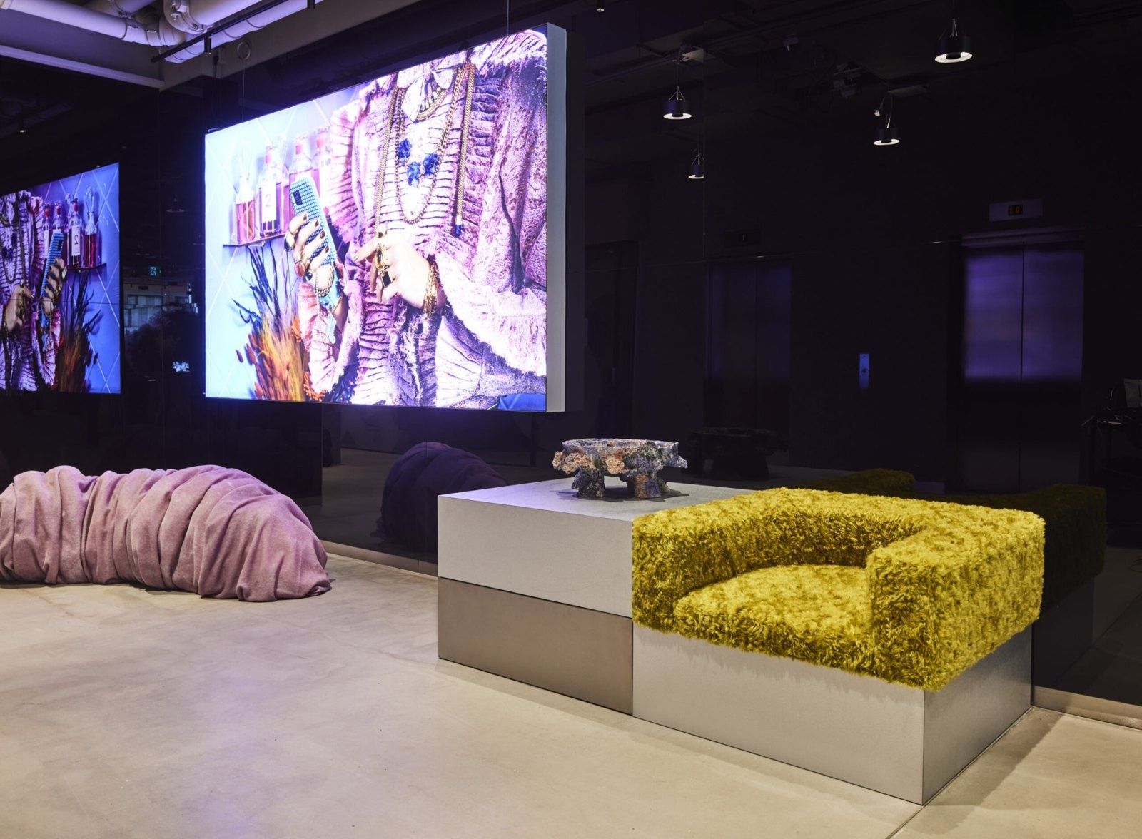Klarna: Dark lounge with a large video wall, a yellow textured armchair on a grey block and a pink soft sculpture