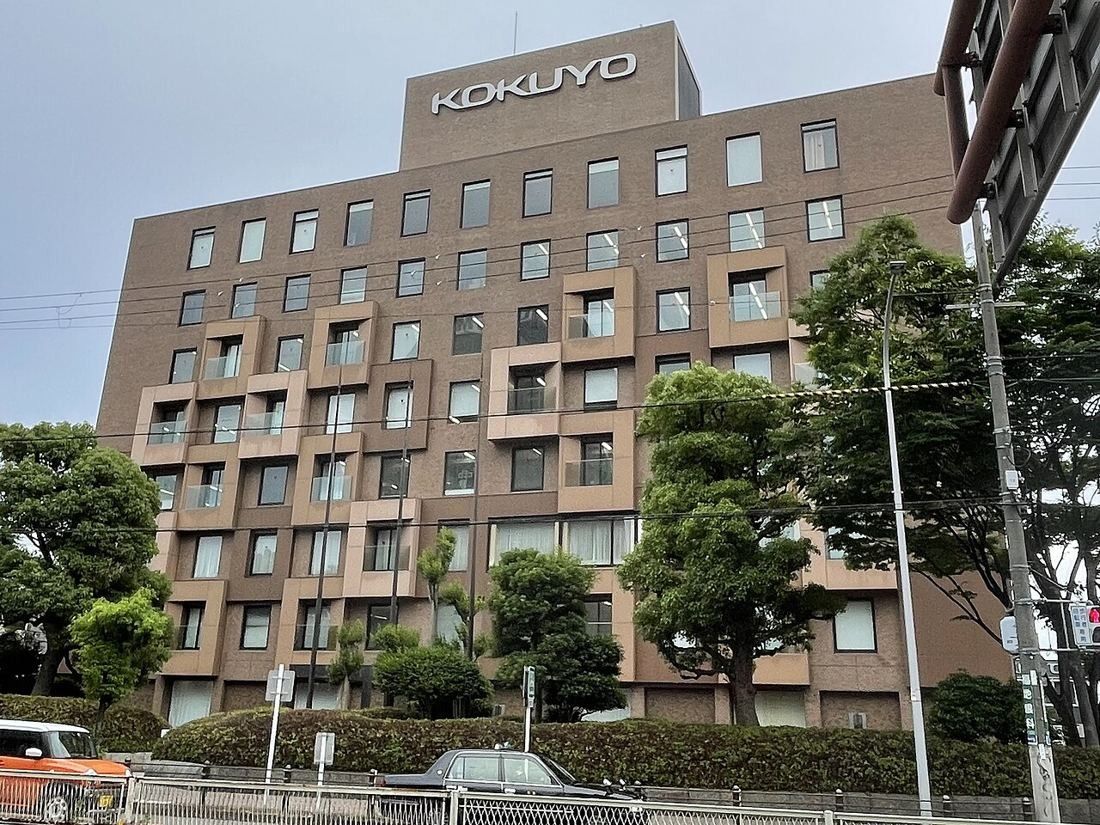 Brown nine-storey office building with the KOKUYO name on the roof, behind street trees