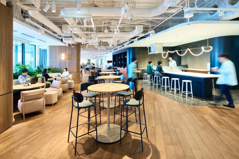 KOKUYO: Café area with high tables, armchairs and a long counter under exposed ceiling ducts