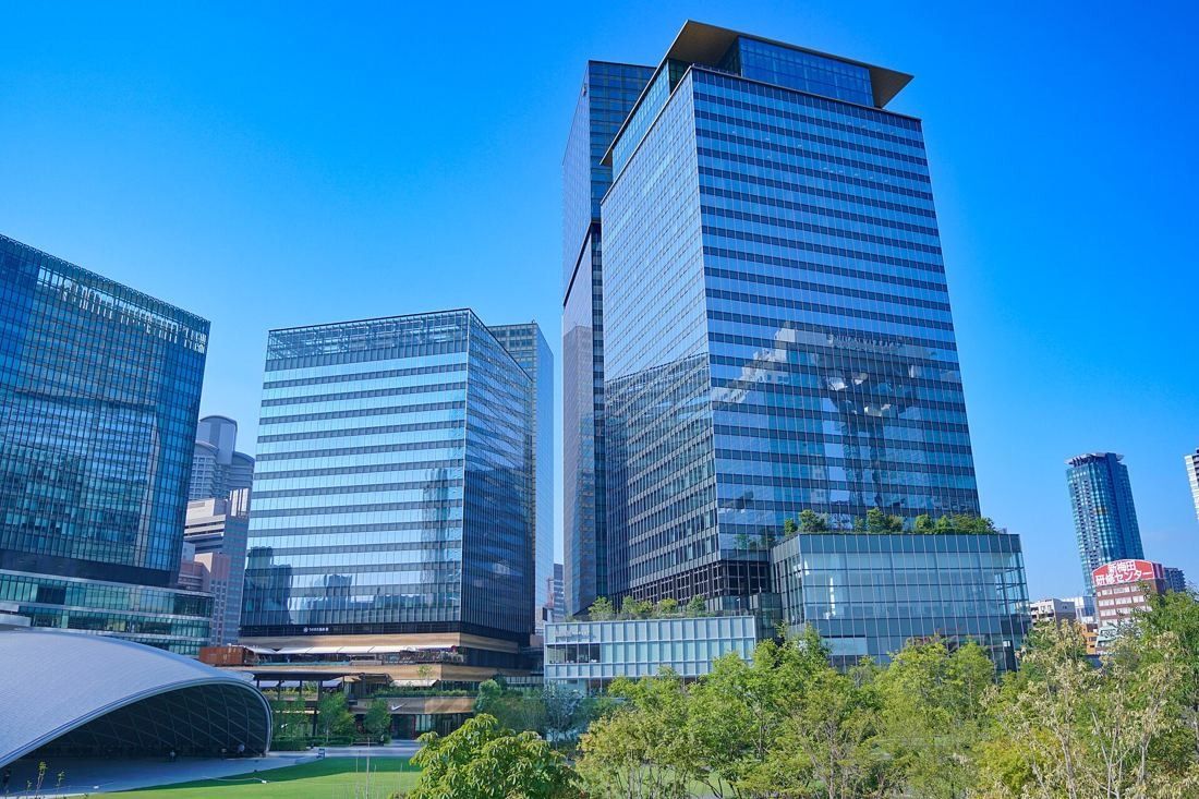 Glass office tower rising above greenery and a curved white roof under a deep blue sky