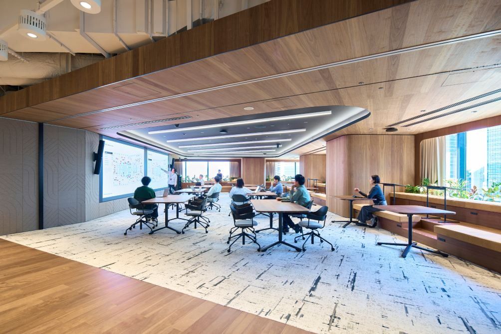 KOKUYO: Room with round meeting tables on a patterned rug, wooden ceiling and wide windows