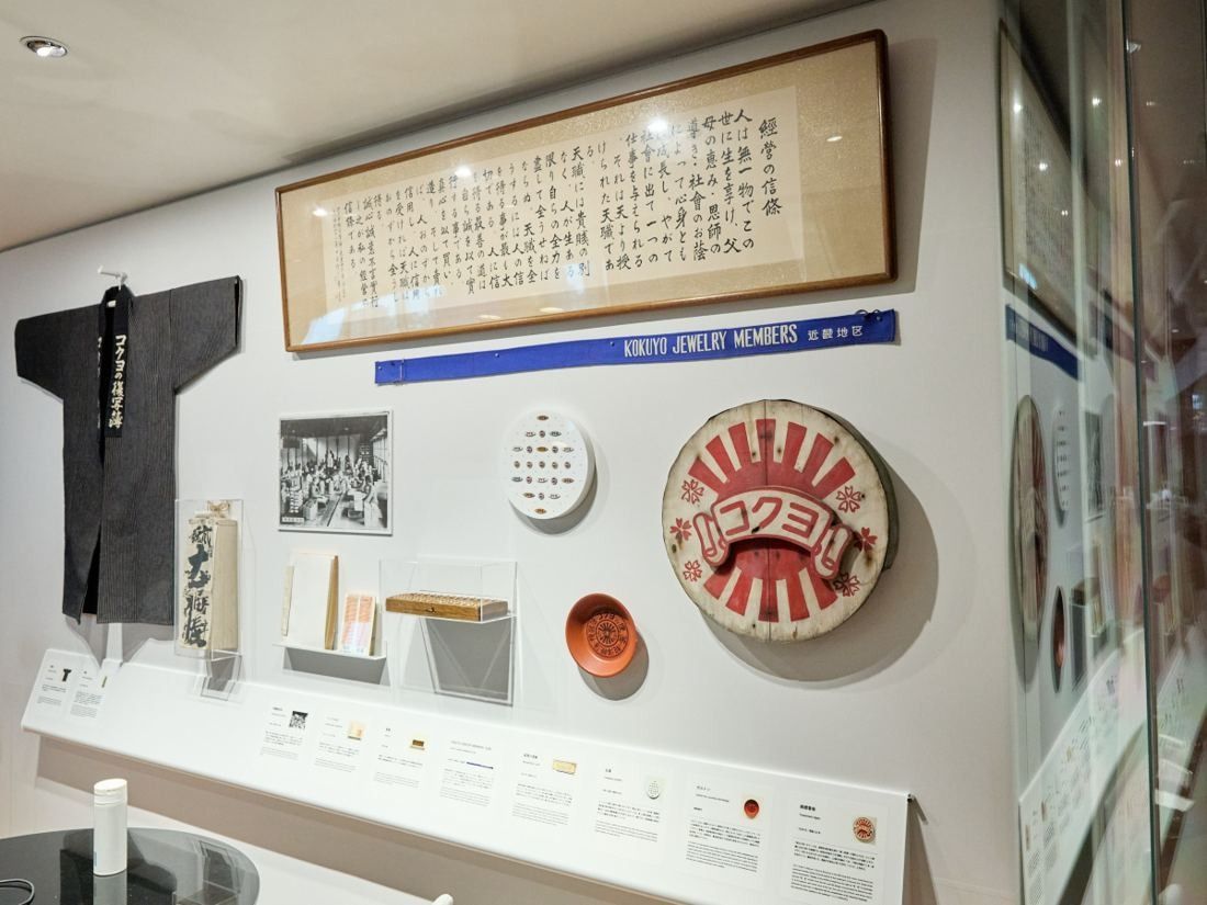 KOKUYO: Wall display with a framed calligraphy, a dark happi coat, an old round sign and small objects