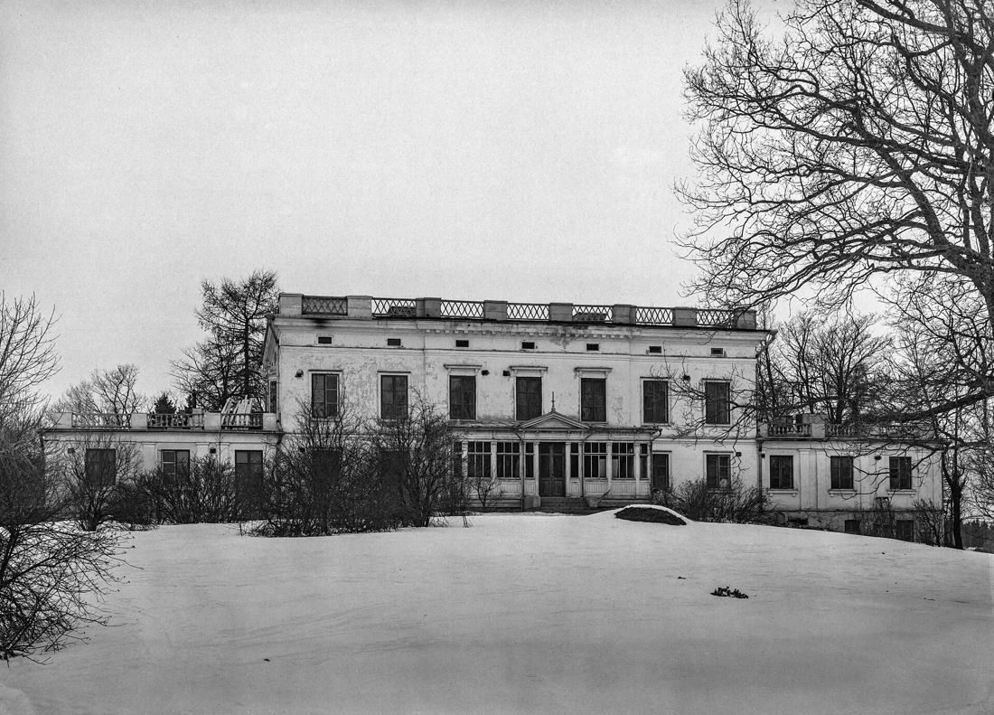 Black-and-white view of a long manor house beyond a snowy field and bare trees