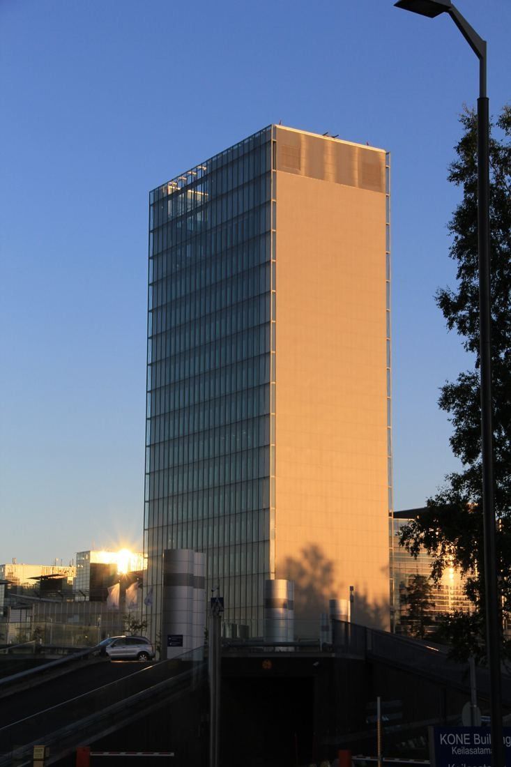 KONE: Glass tower lit orange by the low sun behind trees and a lamppost