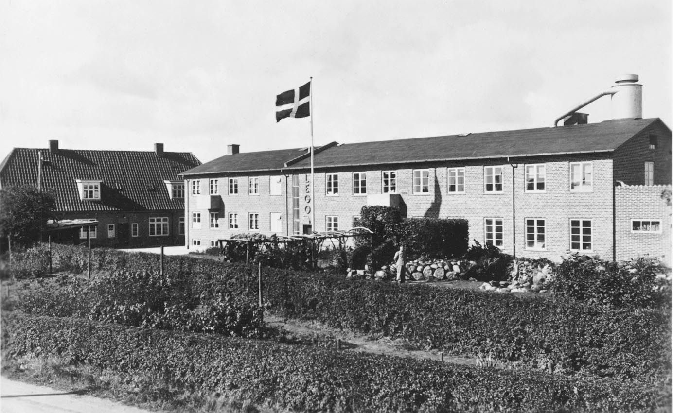 Black-and-white photo of a two-storey factory with a Danish flag and a garden in front