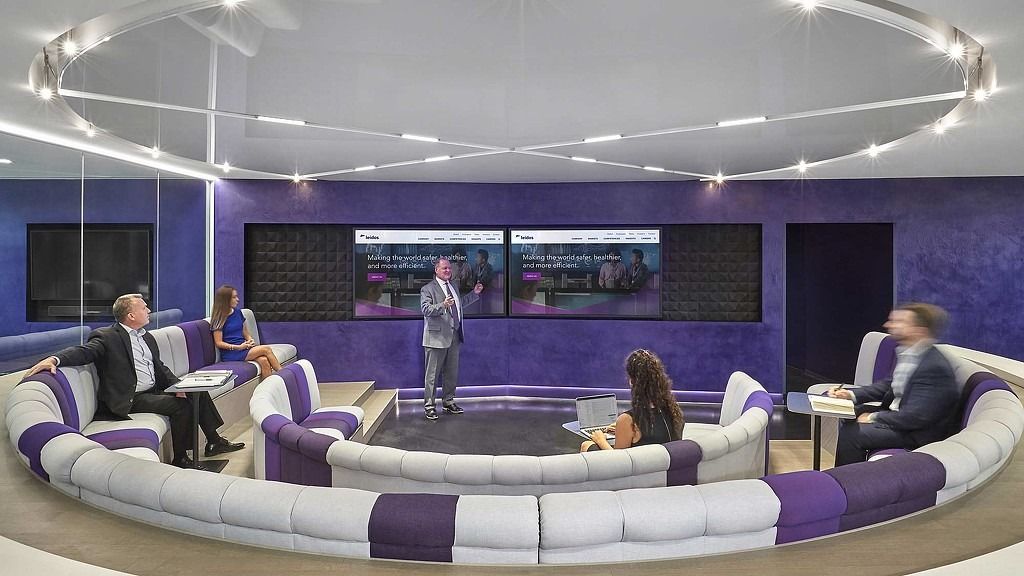 Leidos: A round sunken lounge with white curved sofas and purple cushions below a ring of light