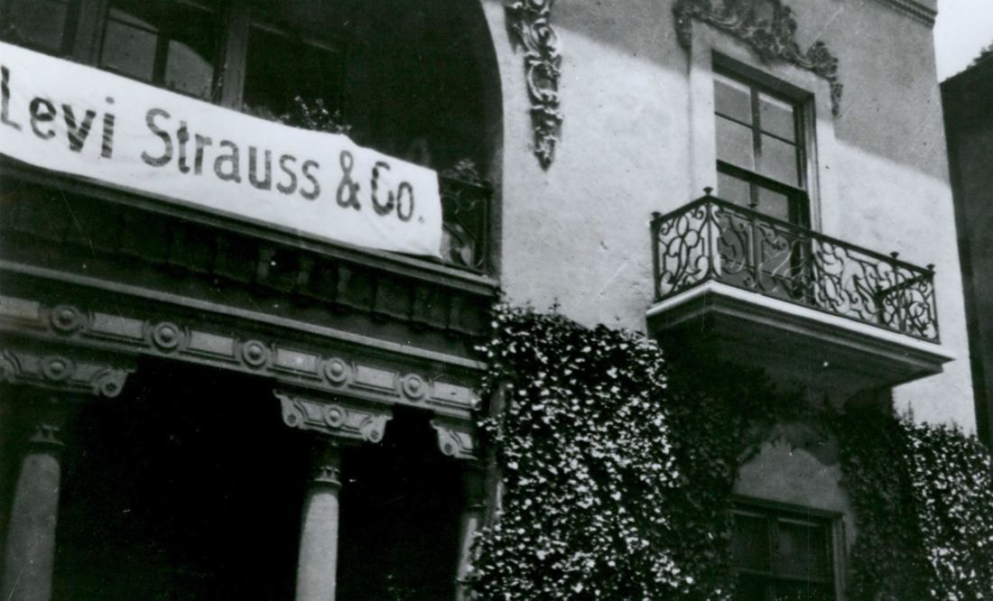 Black-and-white photo of a house facade with ornate balcony and ivy, a banner reading Levi Strauss & Co. hung across the porch