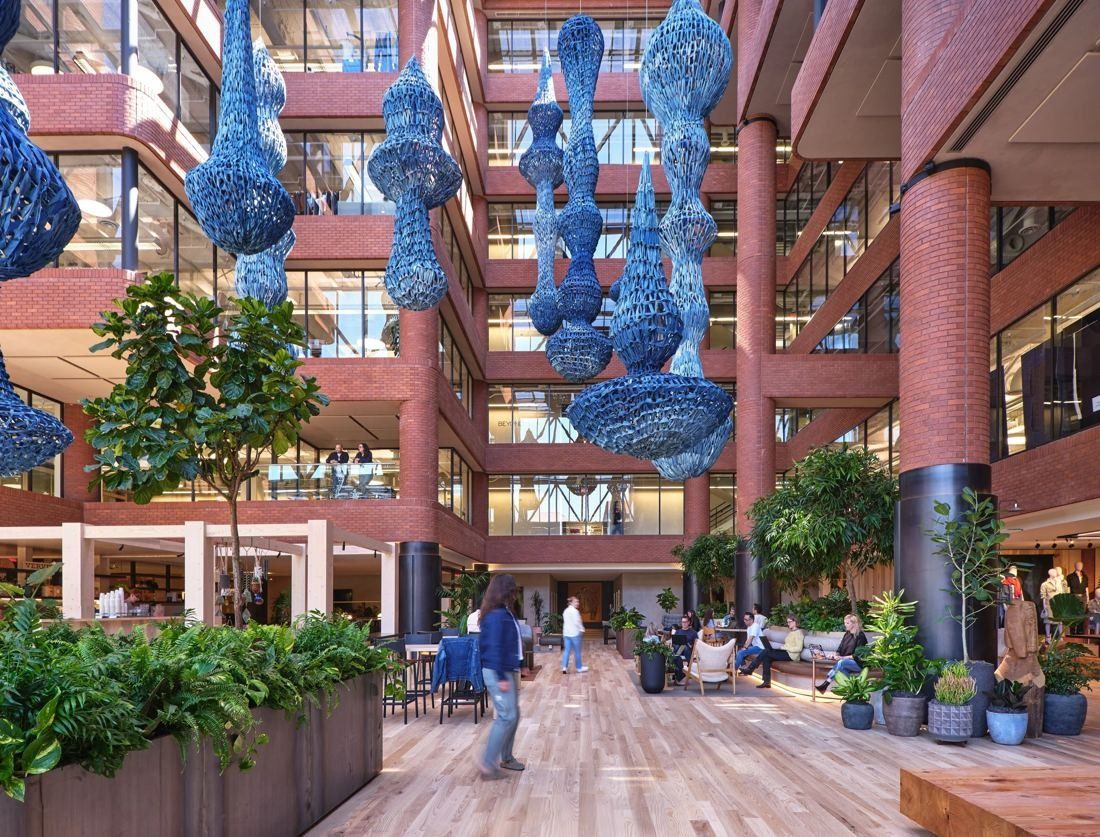 Tall brick atrium with glass-fronted office floors and large blue woven sculptures hanging from the skylight over a wood floor with plants