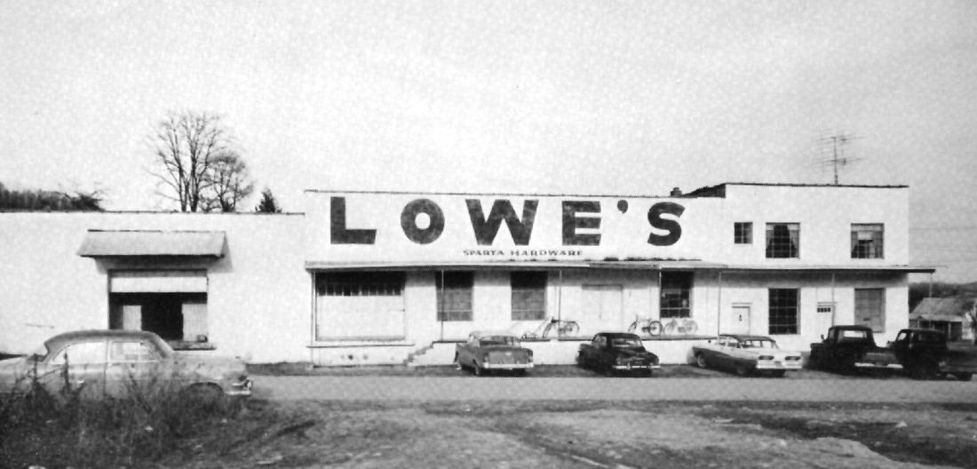 Black-and-white photo of a low store building with large LOWE'S lettering and parked cars in front