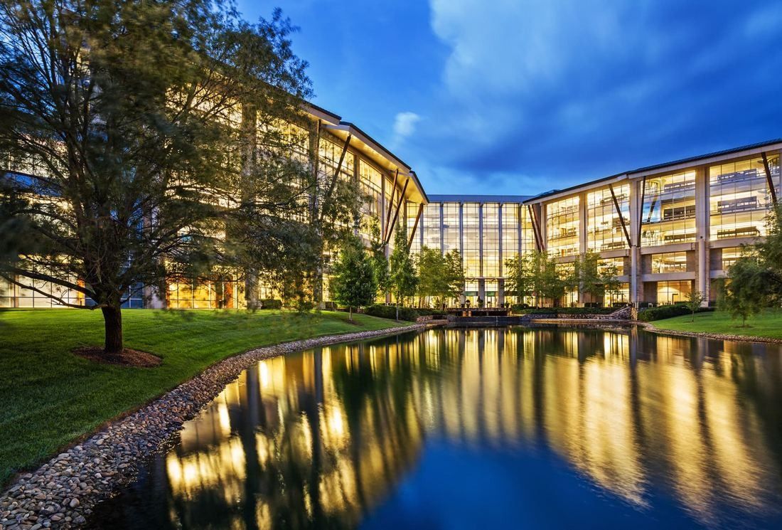 Curved glass office buildings lit at dusk, reflected in a pond surrounded by lawn and trees
