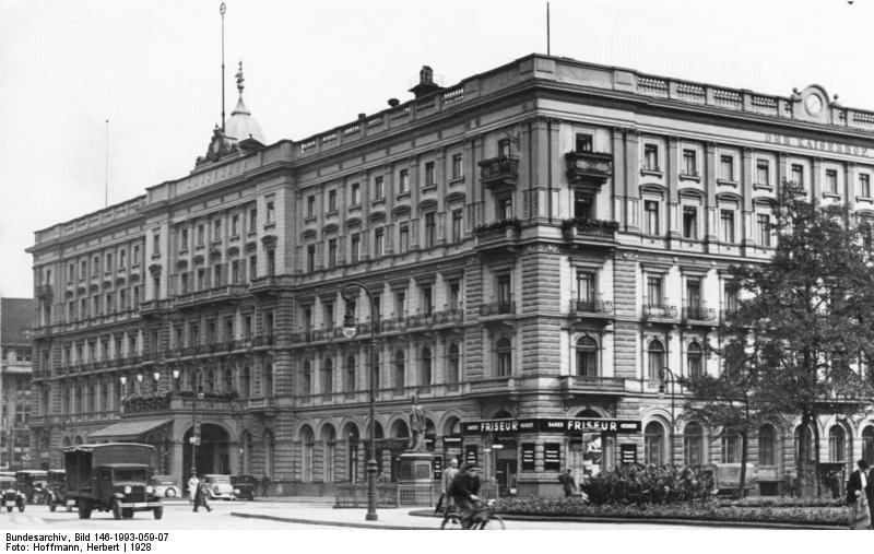 Black-and-white photo of a large ornate corner hotel on a square, with old cars in front
